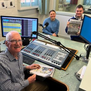 An older man and a young woman and man in their early 20s sit in a radio studio holding newspapers. They are looking at the camera and smiling.