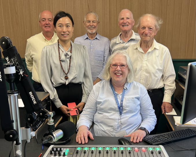 One woman sitting behind a radio panel with 5 people behind her