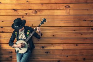 A man playing a banjo wearing a cowboy hat in front of a pine wood wall.